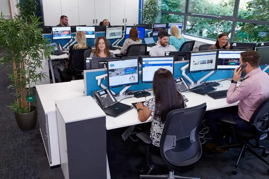 Group of office insurance brokers sat at their desks, in a brightly lit workspace. Some are engaging with computers or phones.