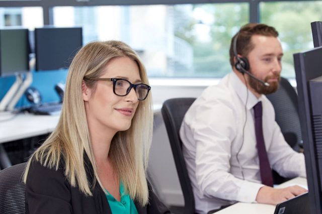 Smiling woman with blonde hair wearing a headset, seated at a desk and looking at a computer screen.