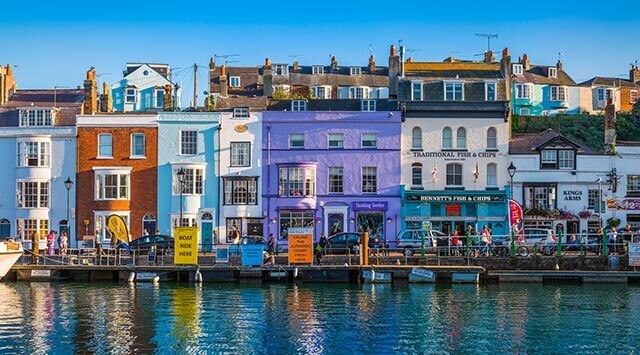 A waterfront scene with colorful buildings reflected in the water. The structures include a purple building, a fish and chips shop, and several pubs. There are people walking along the street under a clear blue sky.