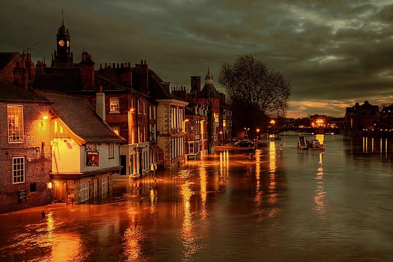 A row of brick buildings submerged by flood at dusk, with street lights under a cloudy sky.