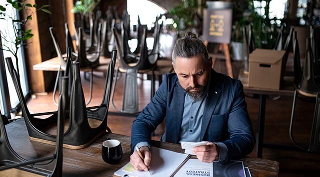 A man with a beard and a man bun is sitting at a table in a dimly lit room. He is reading a piece of paper with a serious expression. There are stacked chairs on tables behind him and a coffee cup on the table in front.