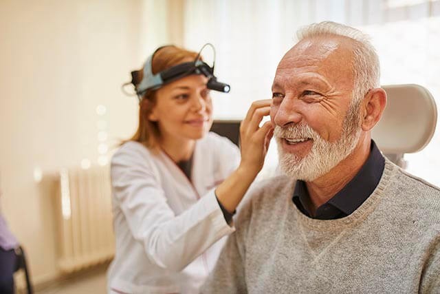 An audiologist examines an older man's ear using a headlamp in a medical office.
