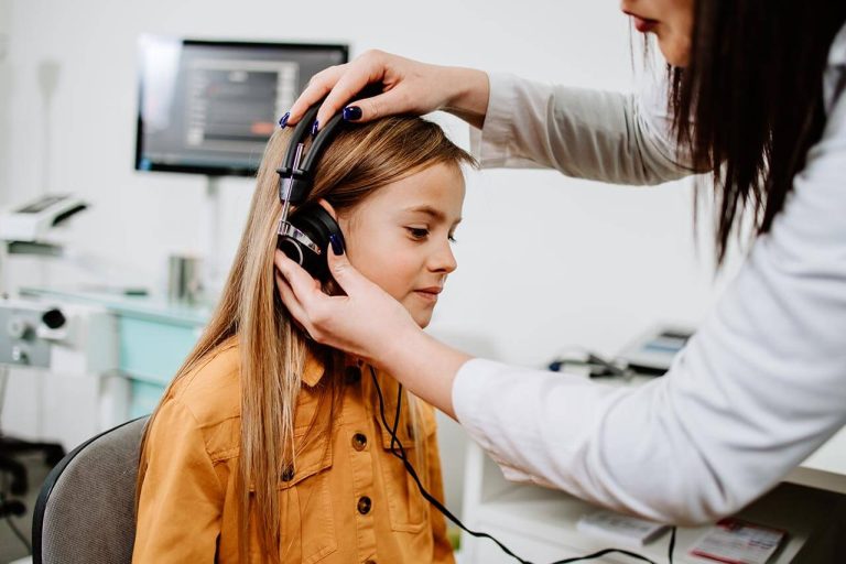 Young girl sitting while a woman adjusts headphones in an audiology office.