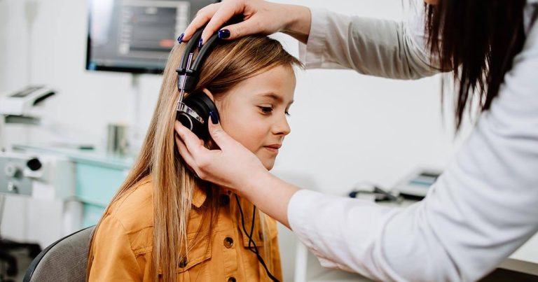 Young girl sitting while a woman adjusts headphones in an audiology office.