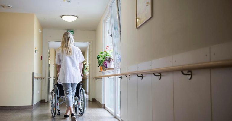 Caregiver walking down a clean, bright hallway, pushing an empty wheelchair.