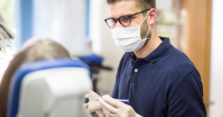 Dentist in navy shirt and mask focused on a patient sitting on a dentist chair.