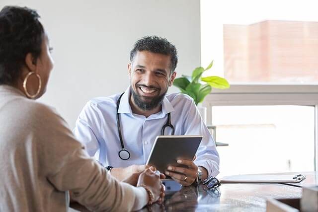 Male doctor smiling while holding a tablet and speaking to a female patient.