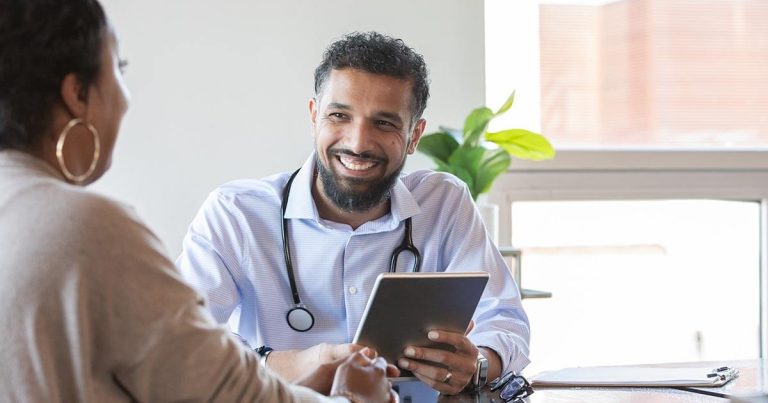 Doctor holding a tablet, listening to a patient in a well-lit consultation room.