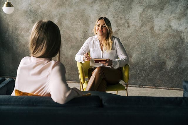 Two women in a therapy session, one holding a notebook and the other seated on a sofa.