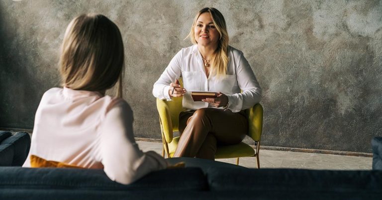 Two people are in a therapy session. Both are seated facing each other. The therapist is holding a pen and notepad.