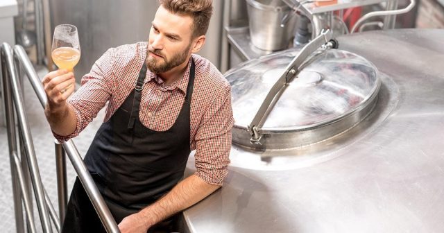 A person wearing an apron inspects a glass of beer next to a large brewing tank with more brewery equipment around him.