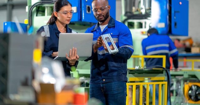 In a bustling factory setting, two individuals discuss both the intricate metal part held by the one in safety glasses and blue jacket and explore business insurance products on a laptop. Machinery hums in the background, underscoring their meticulous approach to innovation and protection.