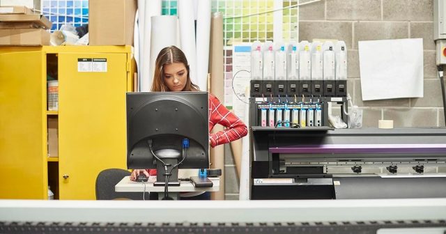 Woman in a plaid shirt standing by a printer in a print shop with printing equipment around her.
