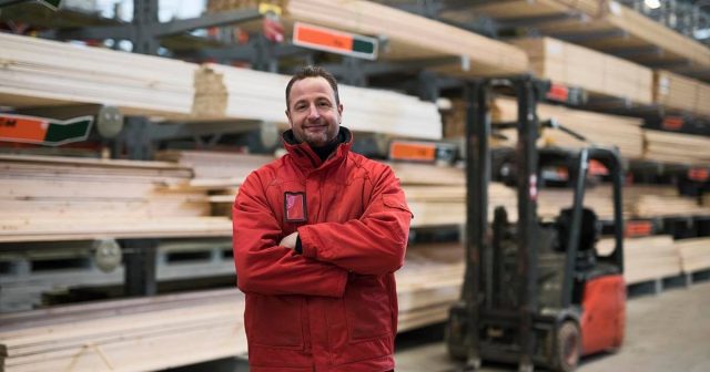 A commercial timber merchant in a red jacket is standing in a lumber warehouse with planks and a forklift.