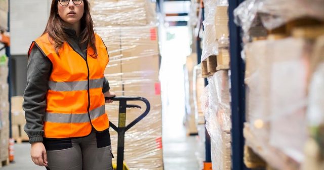 Warehouse delivery worker operating a pallet jack in a well-lit warehouse aisle with stacked boxes.