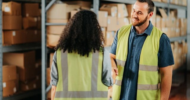 Warehouse staff in high-vis vests shaking hands in front of shelves with boxes.