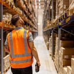 Man in reflective vest walking through a warehouse aisle, holding a device and looking up.