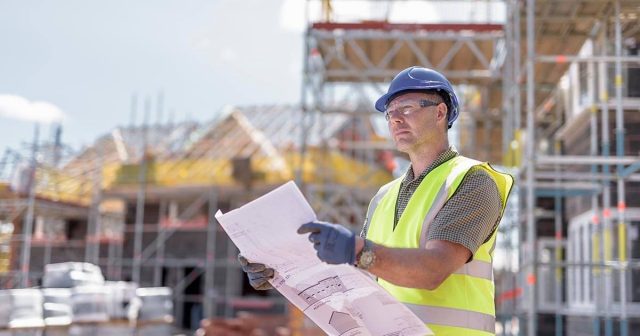 A construction worker, clad in a yellow safety vest, blue hard hat, and gloves, examines blueprints at a bustling site. Scaffolding rises against the clear sky.