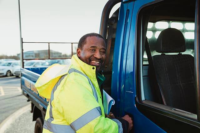 A smiling man in a hi-vis is outside of a blue truck parked in car park.