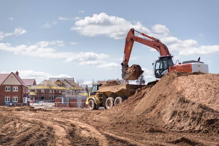 Excavator moving soil at a construction site with residential houses under construction in the background.