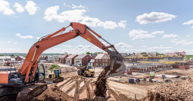 An excavator moves dirt at a construction site with several red brick houses and scaffolding in the background. The sky is clear with a few clouds, and construction vehicles can be seen on the dirt-filled landscape.