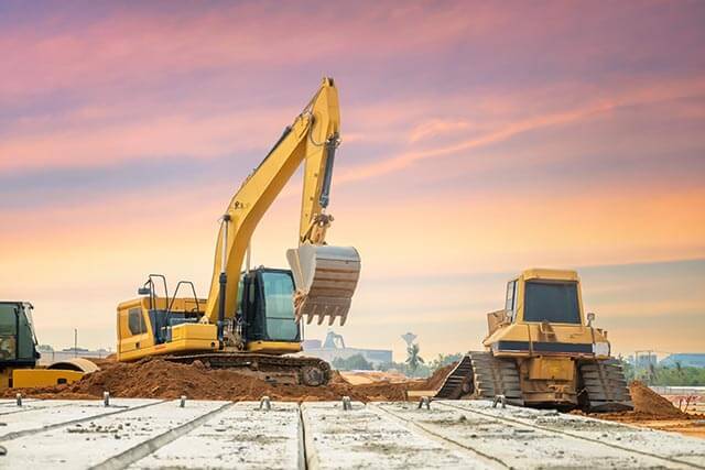 Construction site with an excavator lifting dirt and bulldozer moving, all under a colorful sunset sky with concrete slabs in the foreground.