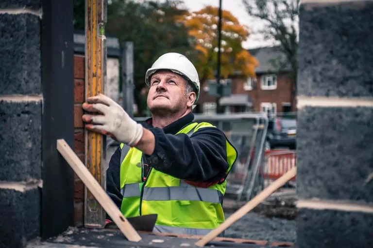 Worker measuring a vertical structure at a construction site with autumn foliage in the background.