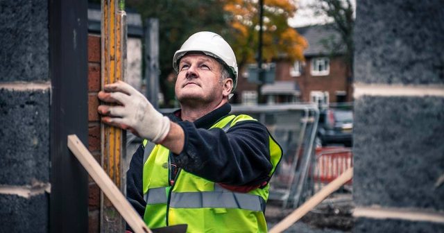 A construction worker in a white hard hat and yellow safety vest holds a measuring instrument against a wall. He is on a construction site with a blurred background of trees and houses.