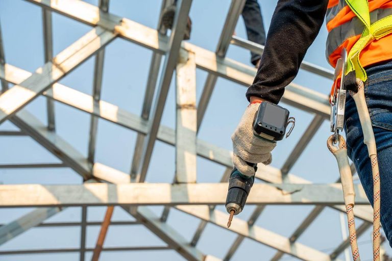 Worker in safety gear holding a drill on a roof, standing on a metal framework under a clear sky.