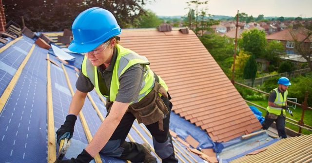 Two construction workers wearing blue helmets and safety vests are installing roofing materials on a house. One worker is in the foreground, actively working, while the other is in the background. Houses and trees are visible in the distance.