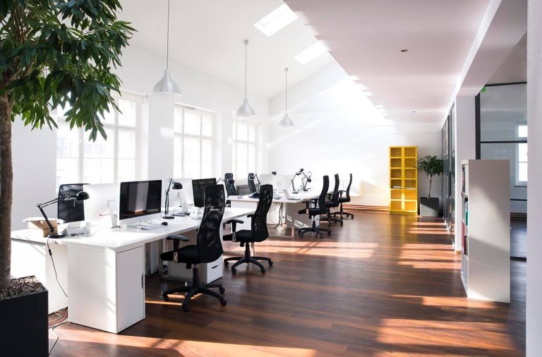 Empty office space with bare desks and chairs, large windows letting in natural light, and plain white walls.