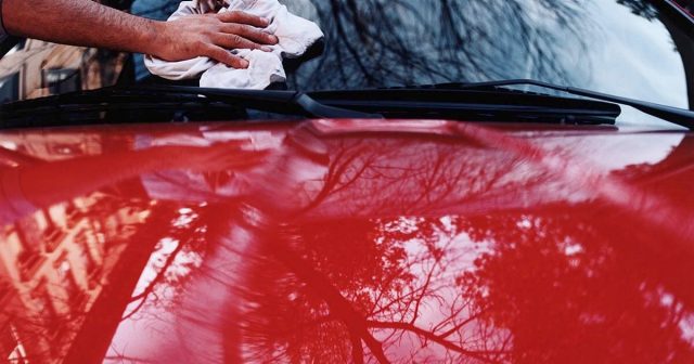Person cleaning the windshield of a red car with reflections of trees and buildings.
