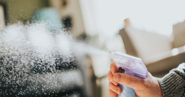 Hand spraying mist from a spray bottle with a blurred background of indoor furniture.