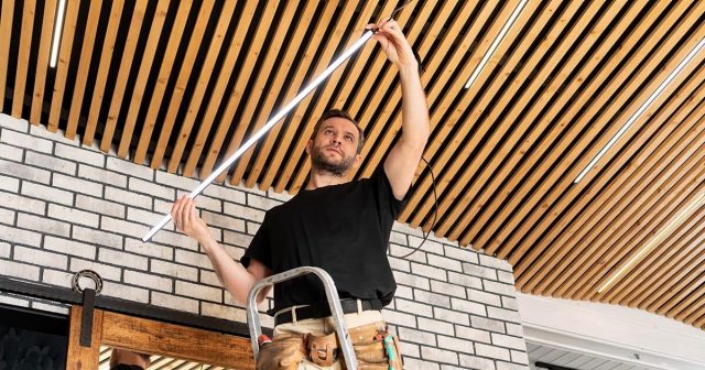 Electrician installing an LED light fixture on a ceiling with wooden slats.