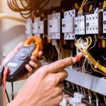 A person uses a digital multimeter to test electrical wires in an open electrical panel. The panel contains various cables, switches, and connectors. The hand is adjusting the wires, with red and black meter probes visible.