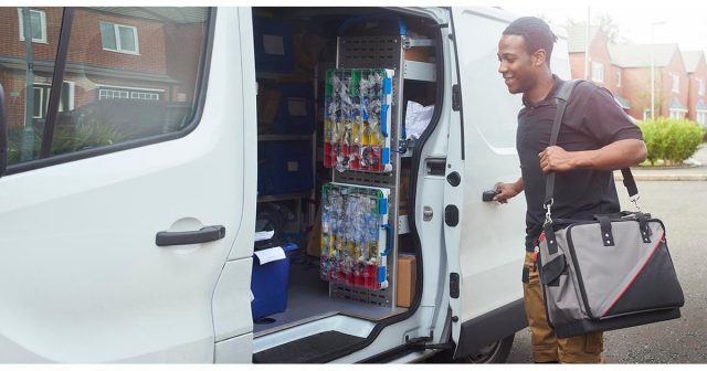 A person in a black shirt carries a gray and black bag while opening the side door of a white van. Inside, shelves are stocked with various supplies. Houses and a pavement are visible in the background.