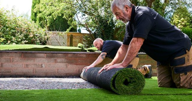 Two men installing artificial grass in a garden, with trees and shrubs in the background.