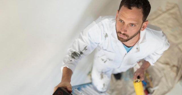 Painter wearing a white jumpsuit on a ladder using a roller and looking upwards.