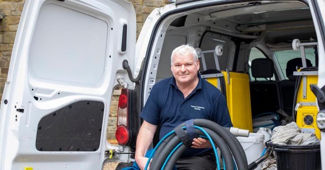 Man sitting at the back of an open white van with cleaning supplies, holding a large hose.