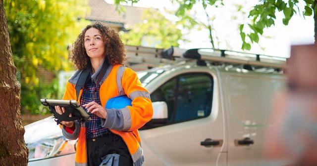 Contractor in safety jacket holding a helmet and a tablet in front of a parked van on a street.