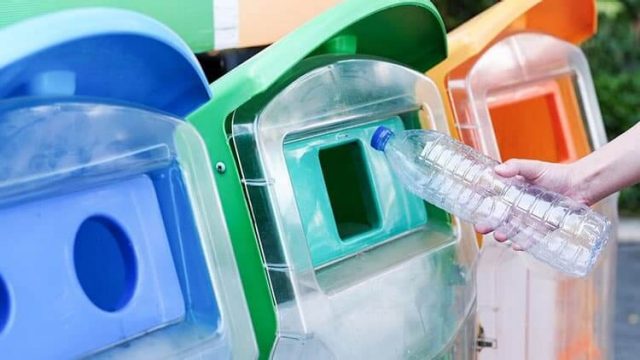 A hand places a plastic bottle into a recycling bin with a blue lid. The bin is situated next to other bins with green and orange lids, likely for different types of recyclables. The background is slightly blurred.