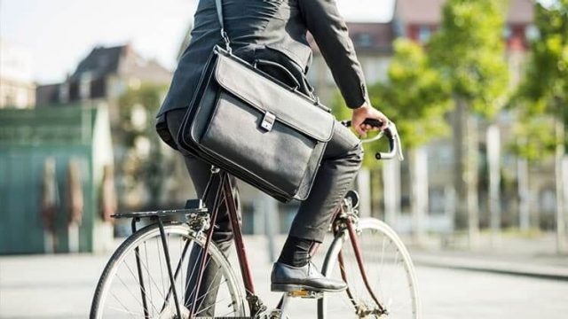 A person in a suit rides a bicycle through a cityscape. They have a black briefcase slung over their shoulder. Buildings and green trees are visible in the blurred background.