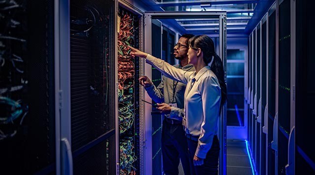 Two individuals stand in a dimly lit server room. They are examining a server rack filled with cables and equipment. One person points at the rack while both appear to be discussing or troubleshooting the setup.