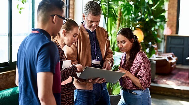 Four people are gathered in an informal setting, looking at a laptop held by an older man. They appear to be discussing or working collaboratively. The background includes large windows and green plants.