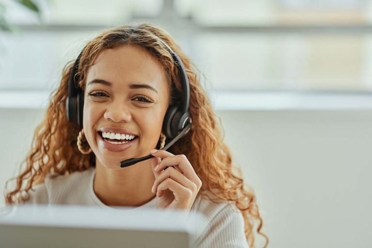 A woman is wearing a headset, smiling during a video or a customer service call in an office setting.