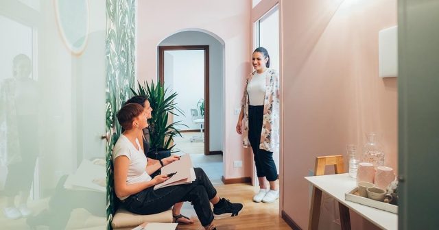 Three people in a hallway, two sitting, one standing and smiling at them near a plant.