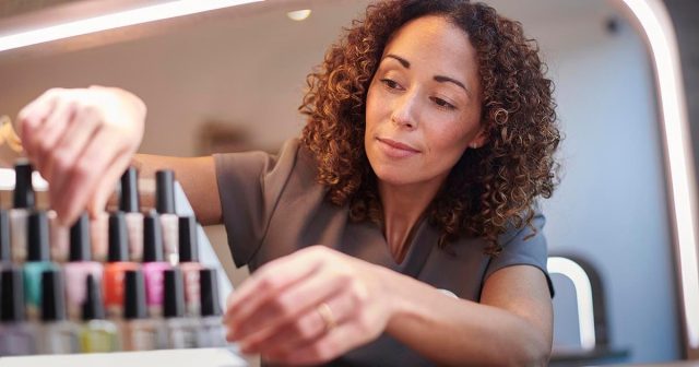 Nail technician sorting bottles of nail polish on a shelf in a salon, with soft lighting.