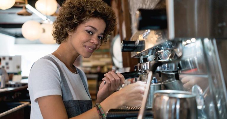 A person with curly hair is operating an espresso machine in a coffee shop. They are wearing a gray apron and smiling at the camera. There are shelves with items and round hanging lights in the background.