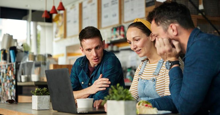 Three people sitting at a café table, looking at a laptop screen and discussing.
