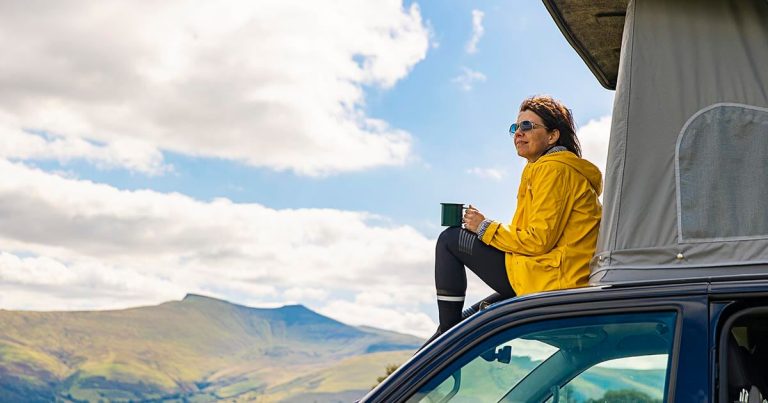 Camper sitting on a vehicle roof, holding a mug, with rolling hills in the background.